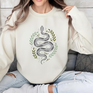 May include: Cream-colored sweatshirt featuring a gray snake design encircled by green leaves. The snake has a detailed pattern and is centered on the front of the shirt. The design is simple and elegant, suitable for casual wear.