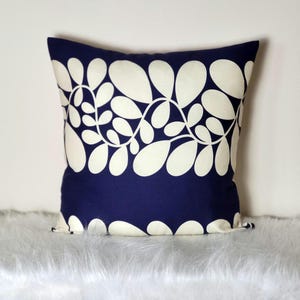 May include: A square cushion featuring a navy blue base and a white floral design. The cushion is adorned with a repeating pattern of stylized leaves. It rests on a fluffy, white surface. The design is symmetrical.