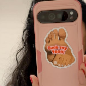 May include: A pink phone case with a graphic of a foot and the text "Suck my toes!". The phone case is held up in front of a person. The person is wearing a black top and a denim jacket.