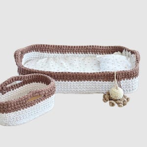 May include: Handmade crochet pet bed set in brown and white. The set includes a rectangular bed with a patterned liner and pillow, and a smaller oval basket. A hanging toy is attached to the bed.