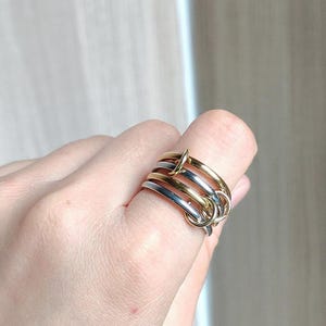 May include: A multi-band ring featuring four interlocking circular bands in alternating gold and silver tones. The design consists of two polished gold-colored bands and two polished silver-colored bands that are linked together to form a single unit.