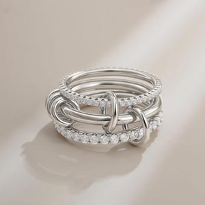 Pode incluir: Um conjunto de anéis de prata com várias bandas entrelaçadas com laços de conexão. Algumas bandas são incrustadas com pequenas pedras preciosas brilhantes. O design é moderno e elegante, com um acabamento polido.