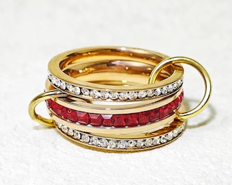 Può includere: Set di anelli dorati con tre fasce impilate. Una fascia presenta una fila di pietre rosse a taglio quadrato, mentre le altre due sono decorate con cristalli trasparenti. Gli anelli sono collegati da anelli dorati.