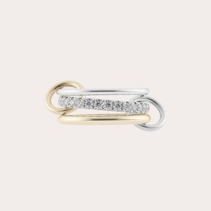 May include: A multi-tone ring featuring a mix of gold, silver, and a band of sparkling stones. The design incorporates interlocking circles, creating a modern and elegant look. The ring is set against a soft, neutral background.