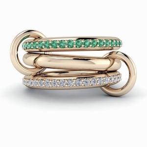Op de afbeelding: Een gouden ringenset met drie in elkaar grijpende banden. Eén band is versierd met kleine, groene edelstenen, een andere met heldere diamanten. Het ontwerp is elegant en modern.