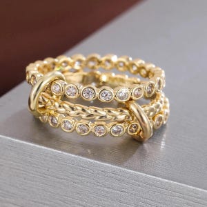May include: A set of three gold-colored rings. Two rings are adorned with small, clear gemstones, while the third ring has a braided design. The rings are interlocked with a gold-colored band.