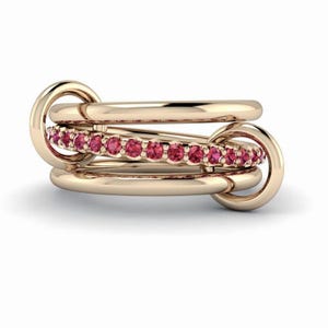 Peut inclure: Une bague dorée avec trois anneaux entrelacés. L'un des anneaux est orné de petites pierres roses rondes. La bague a un design moderne et est posée sur un fond blanc.