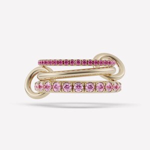 Könnte beinhalten: Ein Goldring bestehend aus drei miteinander verbundenen Bändern. Das obere Band ist mit kleinen rosa Steinen besetzt, das mittlere Band ist glatt poliertes Gold und das untere Band ist mit größeren runden rosa Steinen besetzt, verbunden durch zwei Goldringe.