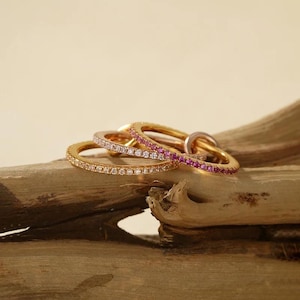 Op de afbeelding: Een verzameling goudkleurige ringen, elk versierd met een rij sprankelende edelstenen. Eén ring heeft heldere stenen, een andere roze en een derde met een mix van roze en heldere edelstenen. De ringen worden gepresenteerd op een stuk drijfhout.