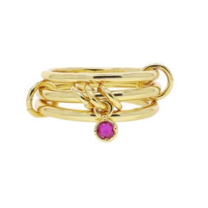 Könnte beinhalten: Goldfarbener Ring mit mehreren durch Glieder verbundenen Bändern. Ein kleiner, runder, magenta-farbener Edelstein hängt in der Mitte. Der Ring hat eine polierte Oberfläche und ein modernes Design.