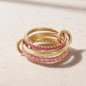 May include: A collection of gold-coloured rings interlinked. Two rings are embellished with pink gemstones, the others are plain. The rings are stacked and connected, creating a unique and stylish jewellery piece.