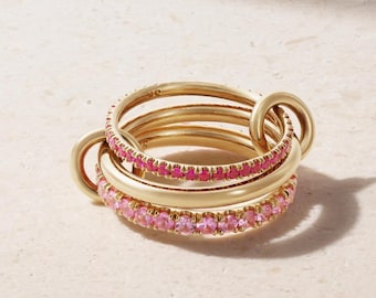 Può includere: Un set di anelli color oro interconnessi. Due anelli sono adornati con pietre preziose rosa, mentre gli altri sono semplici. Gli anelli sono impilati e collegati, creando un gioiello unico ed elegante.