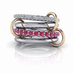 Könnte beinhalten: Ein Stapel silberner Ringe mit goldenen Verbindungsstücken. Ein Ring ist mit kleinen, klaren Steinen besetzt, ein anderer mit kleinen, rosa Steinen. Die Ringe sind ineinander verschlungen und bilden ein einzigartiges Design.