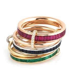 May include: A stack of four metal rings featuring channel-set baguette gemstones in ruby, sapphire, and emerald. One ring is plain rose gold, while another features a vertical bar of small round white diamonds connecting the bands together.
