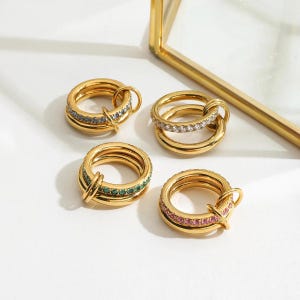May include: Four gold rings with different gemstone details. One ring has blue-grey stones, another has clear stones, a third has green stones, and the last has pink stones. Each ring has a distinctive design with a connecting loop.