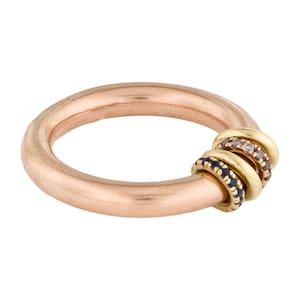 May include: A rose gold ring featuring a thick, smooth band with three smaller, thinner rings attached. One small ring is plain yellow gold, one is set with dark blue stones, and one is set with small, light-colored sparkling stones.