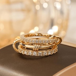 Könnte beinhalten: Ein Stapel goldener Ringe, einer davon mit einem diamantbesetzten Band. Die Ringe sind miteinander verbunden und liegen auf einer dunkelbraunen Oberfläche. Der Hintergrund ist verschwommen mit warmen Goldtönen.