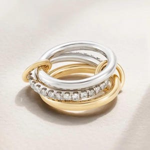 May include: A set of three interlocking rings. One ring is silver, one is gold, and the third is gold with a row of small, clear gemstones. The rings are interlocked, creating a unique and elegant design.