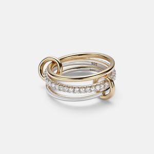 Könnte beinhalten: Ein Set stapelbarer Ringe in Gold und Silber. Ein Ring ist mit kleinen, klaren Edelsteinen besetzt. Die Ringe sind mit goldenen Schlaufen verbunden. Die Ringe sind mit "925" markiert.