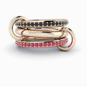 Op de afbeelding: Een stapel van vier in elkaar grijpende roségouden ringen. Eén ring is bezet met zwarte edelstenen, een andere met roze edelstenen en twee zijn effen. De ringen zijn verbonden door een cirkelvormige schakel.