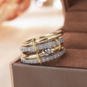May include: A multi-band ring with gold and silver tones. The ring features multiple bands, some encrusted with small, clear gemstones. The bands are connected with gold-colored accents and a twisted silver-colored element.