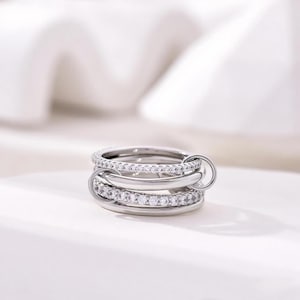 May include: A silver ring with three interlocking bands. Two bands are encrusted with small, clear gemstones. The ring has a modern design and is displayed on a white surface.