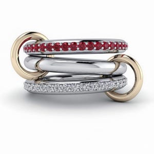 Könnte beinhalten: Ein Stapel von drei Ringen, verbunden durch goldene Verbinder. Der obere Ring ist silberfarben mit einer Reihe roter Edelsteine. Der mittlere Ring ist silberfarben, und der untere Ring ist silberfarben mit einer Reihe klarer Edelsteine. Schmuck.