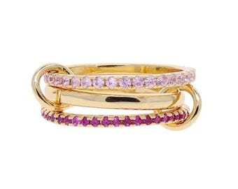 Può includere: Un anello dorato con tre fasce collegate. Le fasce superiore e inferiore sono incastonate con piccole gemme rosa e magenta. La fascia centrale è in oro liscio. L'anello presenta un design unico e interconnesso.