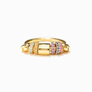 Op de afbeelding: Een gouden ring met een centraal rechthoekig element en kleurrijke edelsteenaccenten. De ring heeft blauwe, gele, paarse en roze stenen, wat zorgt voor een levendig ontwerp. De ring staat tegen een witte achtergrond.