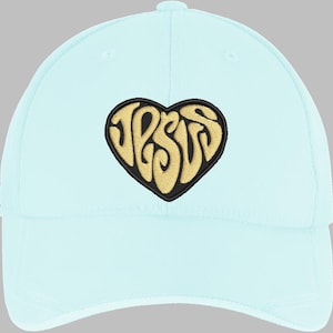 May include: A light blue baseball cap with a curved brim. The cap features a heart-shaped patch with the word "JESUS" embroidered in gold thread, outlined in black. The cap is made of a soft fabric and has a rounded crown.