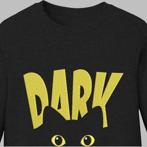 Può includere: Maglietta nera a maniche lunghe con la scritta "DARK" in giallo, con testo stilizzato. Sotto il testo, è visibile la sagoma di un gatto nero, con occhi gialli. La maglietta è di colore melange scuro.
