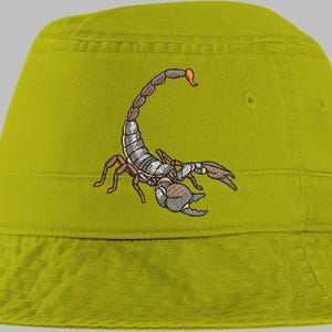 May include: A chartreuse bucket hat featuring an embroidered scorpion design. The scorpion is detailed with brown, gray, and orange accents. The hat has a structured brim and a tonal band around the crown.