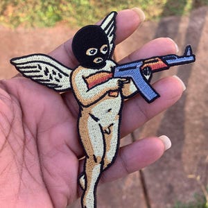 Pode incluir: Patch bordado com uma figura de querubim usando uma balaclava preta e segurando um rifle de assalto azul e laranja. O anjo tem pele bronzeada, asas brancas e é contornado em preto.