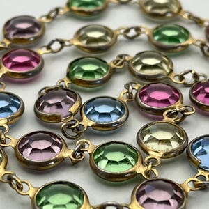 May include: A close-up of a necklace featuring colorful, round gemstones in shades of pink, green, blue, and yellow. The gemstones are set in gold-toned metal settings and linked together with small connecting loops. The necklace is displayed against a white background.