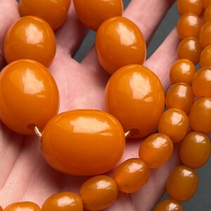 Puede incluir: Un collar vintage de ámbar con grandes cuentas pulidas de diferentes tamaños. Las cuentas tienen un tono naranja cálido y están ensartadas con un cordón delicado. El collar se muestra sobre un fondo neutro, resaltando el color y la textura del ámbar.