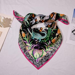 May include: A square silk scarf featuring a black background with a vibrant jungle-themed print including a light blue crocodile, orange pineapples, and green foliage. The scarf is finished with a bright pink border and a geometric grey pattern.