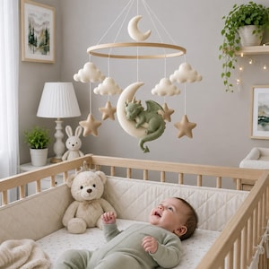 May include: A nursery mobile featuring a central sage green dragon sleeping on a cream-colored crescent moon, surrounded by hanging felt clouds and tan stars suspended from a circular wooden frame with a small moon at the very top center.