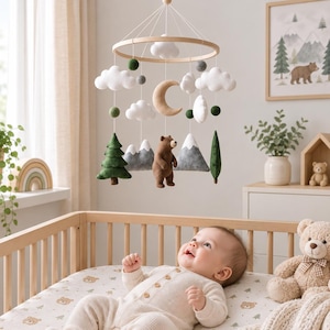 Op de afbeelding: Een babymobiel met een rond houten frame en hangende vilten elementen, waaronder witte wolken, groene dennenbomen, grijze bergen, een bruine beer, een halve maan en kleine groene en grijze vilten balletjes aan witte koordjes.