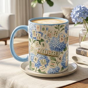Puede incluir: Una taza de café de cerámica con asa y borde azul claro, decorada con un estampado de flores de hortensia azules y fichas de Mahjong. La taza está sobre un platillo pequeño y redondo en una mesa de madera. La taza está llena de una bebida marrón claro.