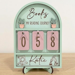 May include: A mint green wooden book counter with the words "Books My Reading Journey" at the top. It has three pink number cards displaying "058". The name "Katie" is written at the bottom, along with decorative illustrations.