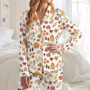Puede incluir: Un conjunto de pijama de camisa de manga larga con botones y pantalón a juego en color blanco, con un estampado integral de calabazas naranjas, peras amarillas, manzanas rojas, setas marrones y flores y hojas en tonos otoñales con ribetes naranjas.