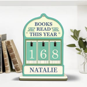 Könnte beinhalten: Ein türkisfarbener und cremefarbener Bücherzähler aus Holz mit dem Text "BOOKS READ THIS YEAR" und der Zahl "168". Der Name "NATALIE" wird unten angezeigt. Der Zähler steht auf einem weißen Regal mit Büchern im Hintergrund.
