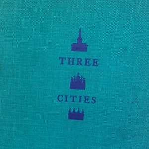 May include: A teal blue book cover with the title "THREE CITIES" printed in dark blue letters. The title is centered on the cover and is surrounded by three stylized images of buildings.