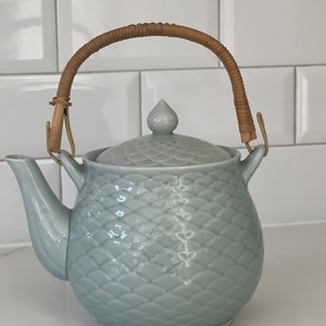 May include: A light blue ceramic teapot with a woven bamboo handle and a textured, scale-like design. The teapot has a lid with a small knob on top.