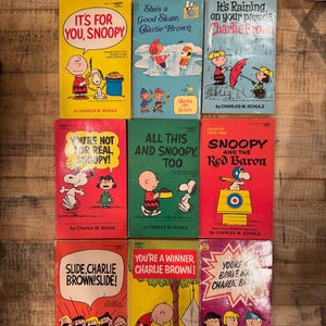May include: A collection of vintage Peanuts comic books by Charles M. Schulz. The books feature colorful covers with titles like "It's For You, Snoopy" and "You're a Winner, Charlie Brown!" The books are arranged on a wooden surface.
