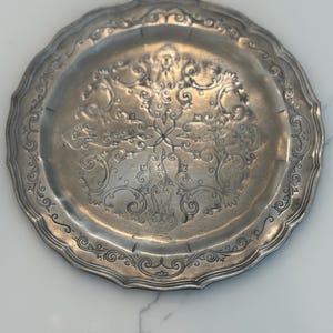 May include: A silver-colored, round tray with a scalloped edge. The tray features an ornate, embossed floral design in the center and along the rim. The tray's surface has a reflective quality, suggesting it is made of metal.