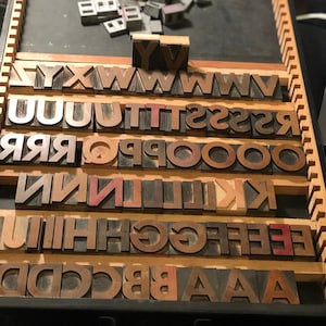 May include: A wooden printing block set with individual letters in a drawer. The letters are arranged in alphabetical order, starting with A and ending with Y. The letters are made of wood and have a dark brown finish.