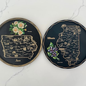 May include: Two black metal trays with gold trim, each featuring a map of a US state. The tray on the left shows Iowa with the text "Iowa" and "Wild Rose". The tray on the right shows Illinois with the text "Illinois" and "Purple Violet".