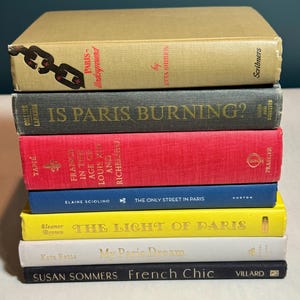 May include: A stack of seven books with titles related to Paris. The books have various colors, including tan, red, blue, yellow, white, and black. Titles include "Paris Underground," "Is Paris Burning?" and "French Chic."