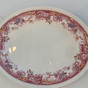 May include: Oval serving platter with a white center and a decorative border. The border features a maroon floral and scroll design with accents of yellow and blue. The edge has a textured, ridged pattern.